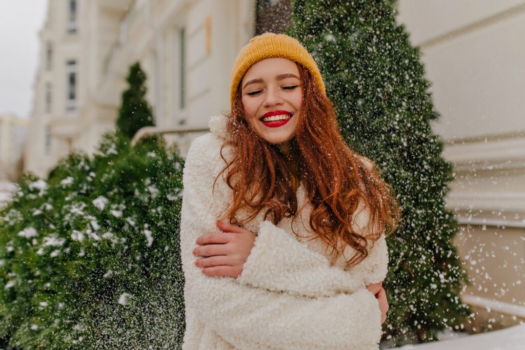 Cheerful ginger lady posing under snow. Smiling lovable woman in knitted hat standing near spruce.